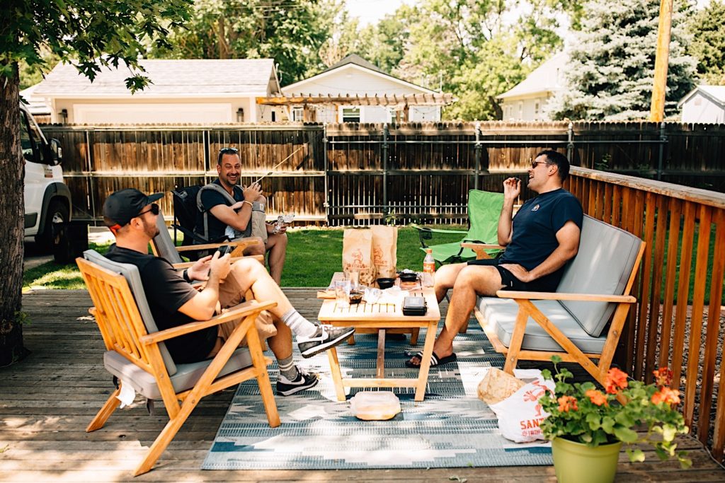 Three men sit on an outdoor patio together with food on the table in front of them while they all smile and laugh