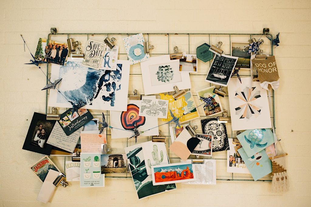 A collage board on a wall consisting of art, postcards, wedding invites, pictures and other items.