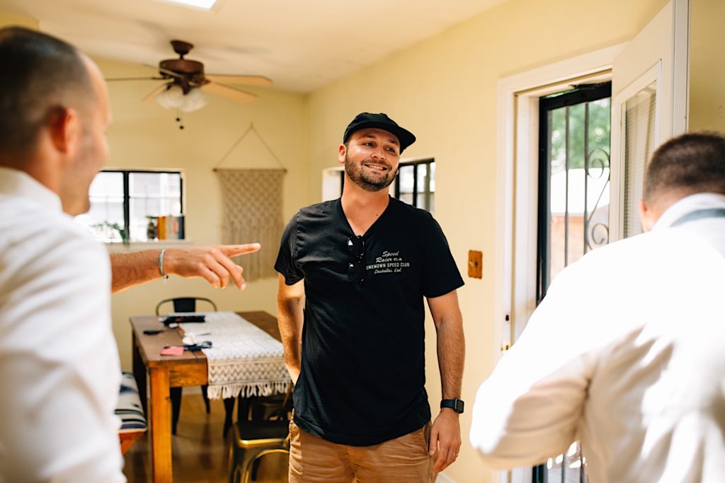 A man standing and smiling at his friends standing on either side of him, one of his friends has his finger pointed to the right