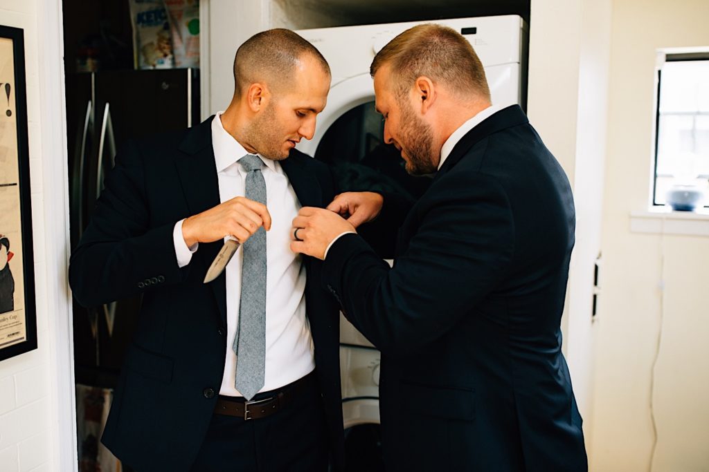 A groom has his friend help to adjust his coat in the laundry room of his home before his wedding