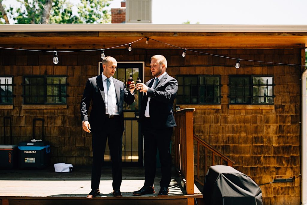 A groom and his friend wearing suits stand outside a home on the back deck with string lights above them as they toast their beers