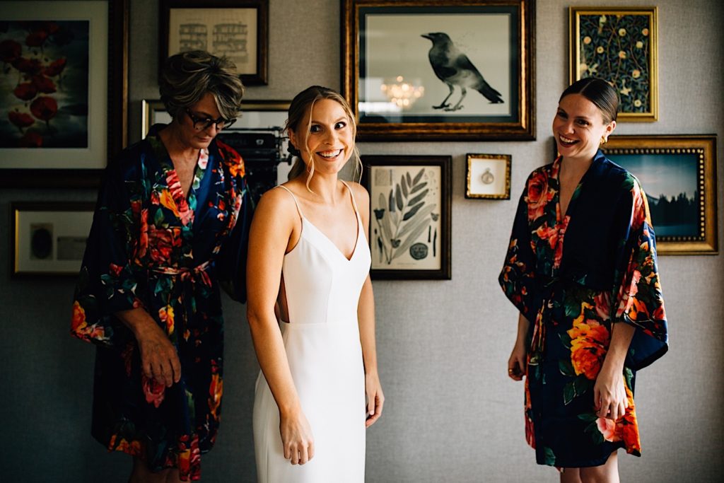 Bride smiles at something behind the camera wearing her wedding dress, there's a woman on either side of her both wearing matching floral robes, photos are hanging on the wall in the background