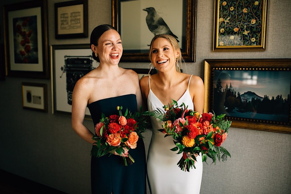 Bride and bridesmaid stand next to each other wearing their dresses, they're each holding bouquets and laughing with one another in a room with framed pictures behind them