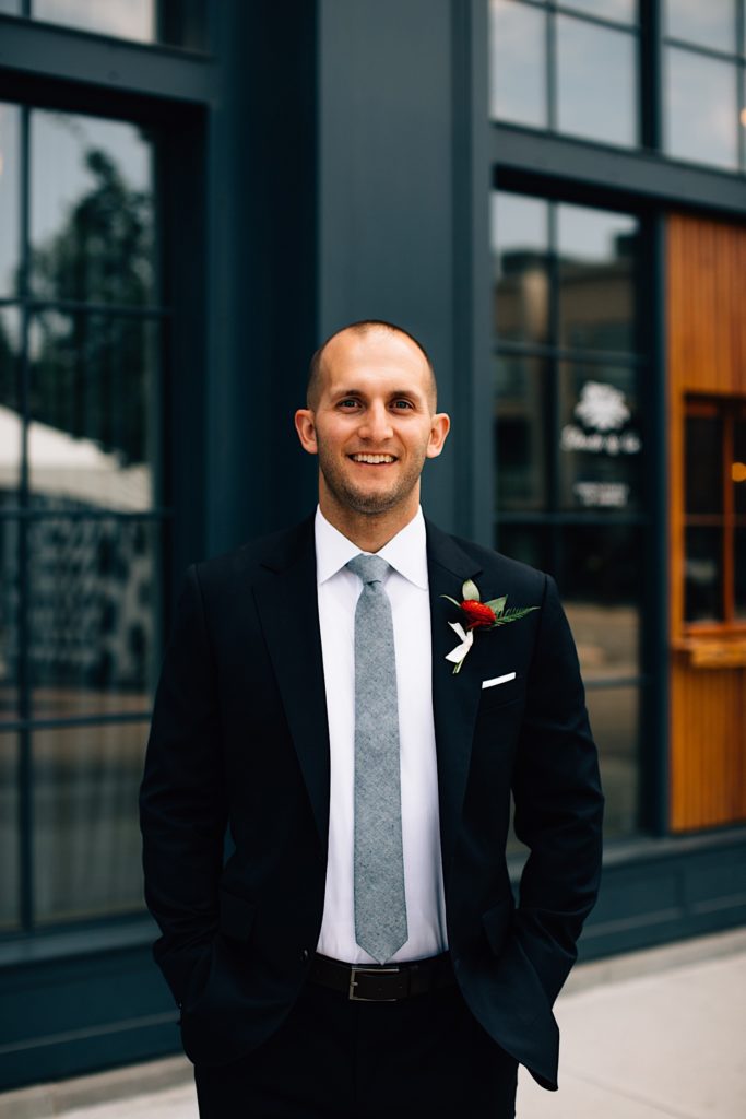 Man wearing a suit and tie with a red flower smiles at the camera in front of a building with lots of windows before his wedding