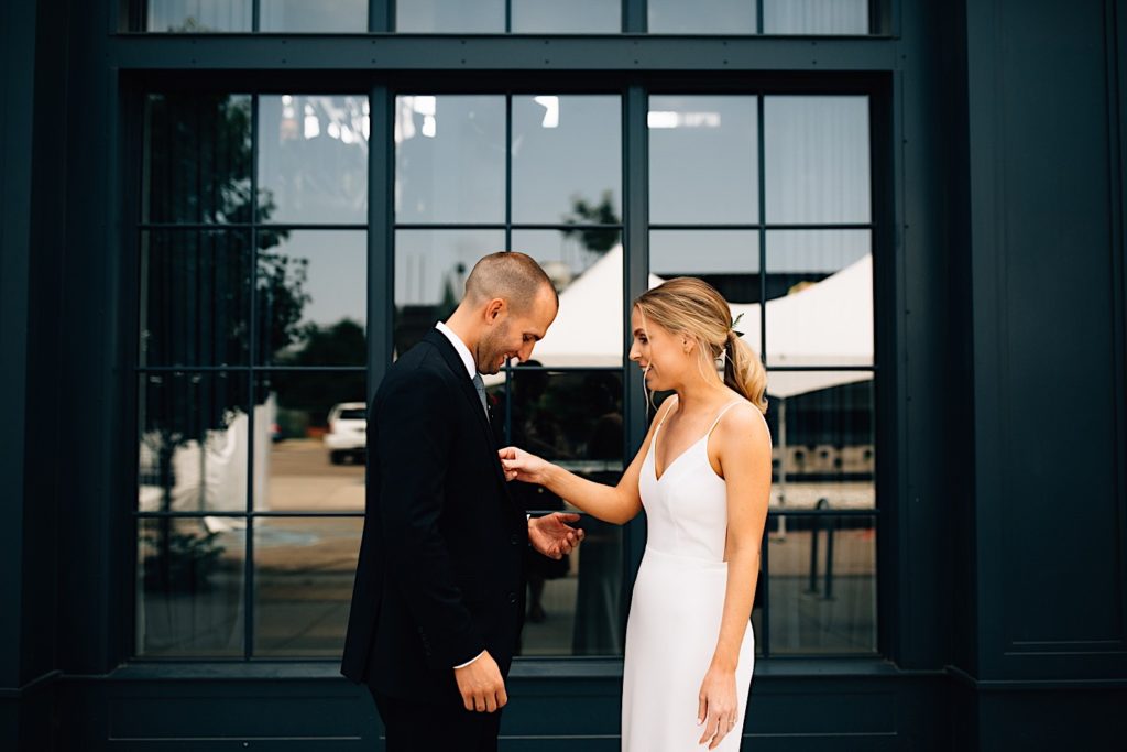 A bride and groom stand together looking at one another, the groom is smiling and looking down as the bride adjusts his tie and smiles