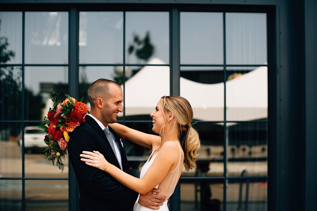 A bride and groom smile at one another and embrace before their wedding, they're wearing their wedding attire and the bride is holding a flower bouquet