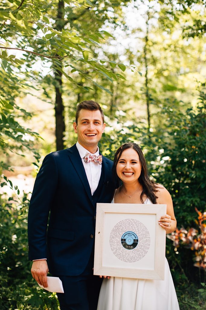 Bride and groom pose and smile at camera, the bride is holding framed artwork from the groom