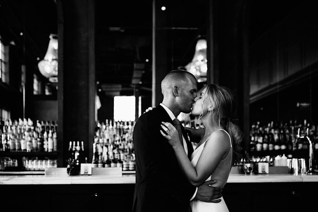 Black and white photo of a bride and groom wearing their wedding attire kissing one another in front of a bar