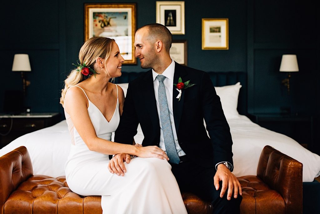 A bride and groom sit on a leather seat in front of a bed, they face one another and smile as they look at one another wearing their wedding attire