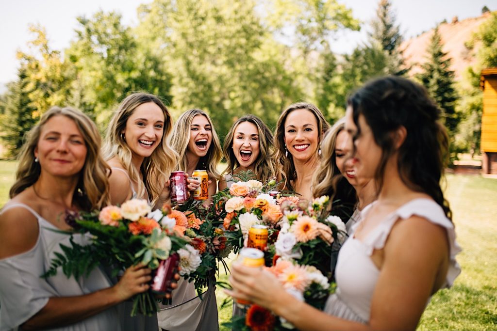 Bride and bridesmaids gather around each other while holding flower bouquets and sodas while smiling and laughing