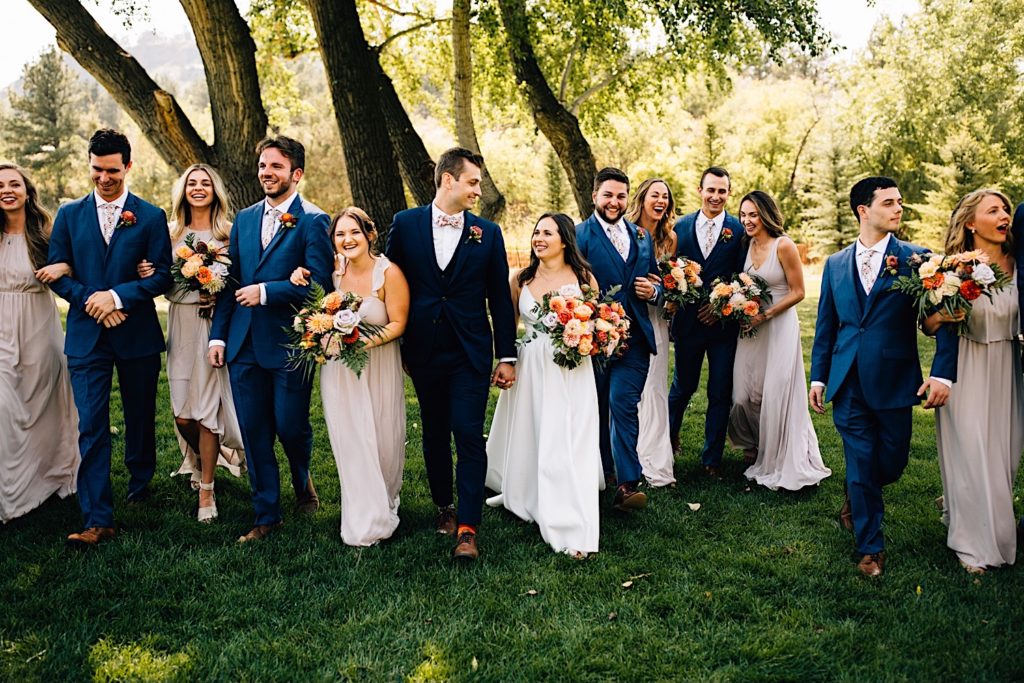 Bride, groom and their wedding party all walk towards camera while holding hands and flower bouquets