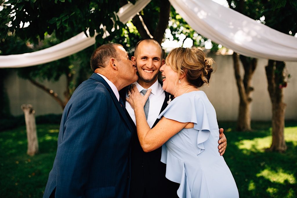 A groom stands in between his parents, his father is about to kiss his cheek and his mother is laughing, the groom is smiling and is dressed for his wedding