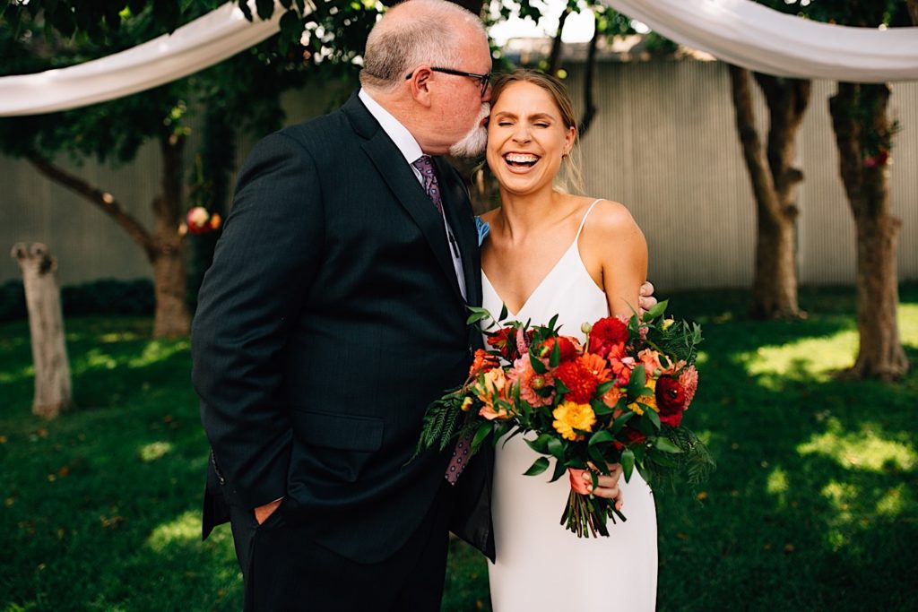 Bride and her father stand next to one another outdoors at their wedding venue, the father kisses his daughter on the temple as she laughs