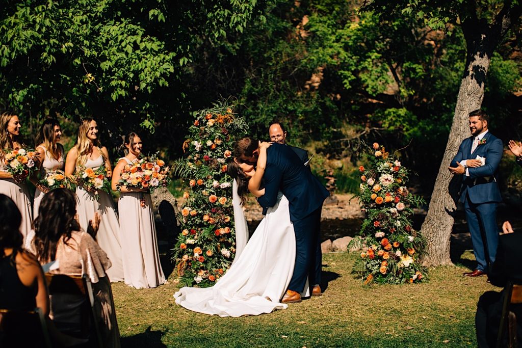 Bride and groom kiss at their Planet Bluegrass ceremony space as their pastor and wedding party watch and celebrate