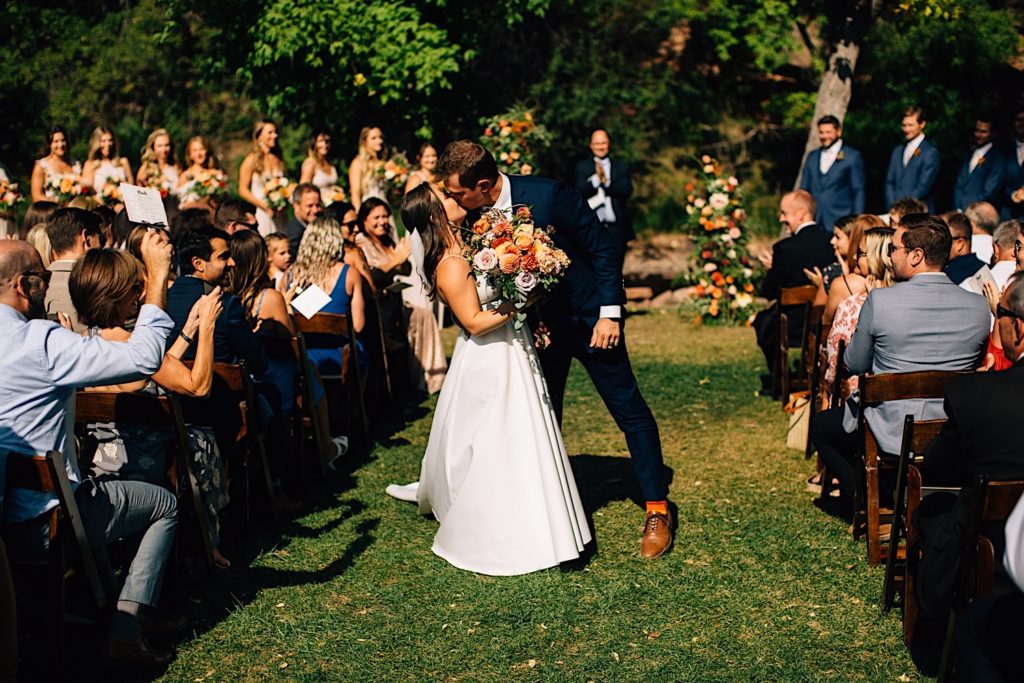 Bride and groom kiss halfway down the aisle exiting their Planet Bluegrass wedding ceremony while their guests watch and clap