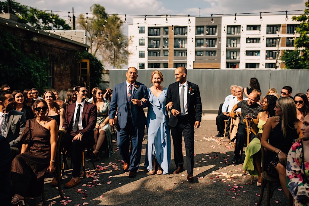 A groom walks down the aisle of his wedding with his mother and father, the mother is between her son and husband and has her arms locked with theirs