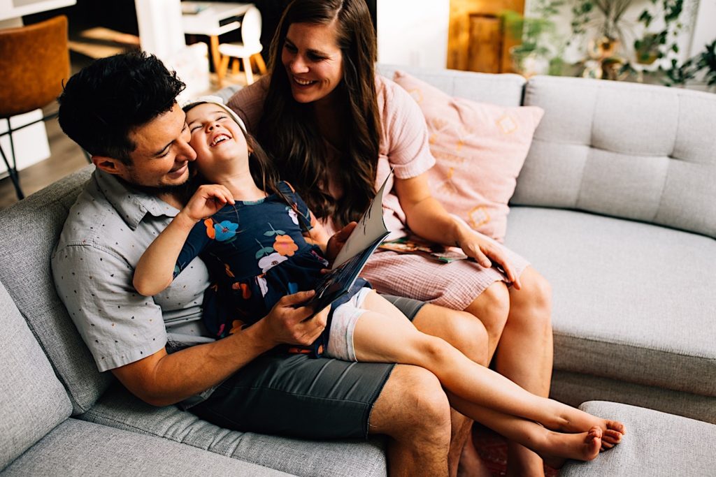 A father sits on a couch with his laughing daughter on his lap while his wife sits next to them.