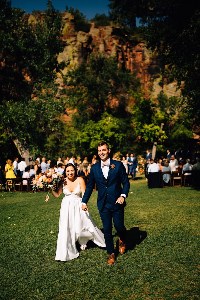 Bride and groom smile while holding hands exiting their Planet Bluegrass wedding ceremony with their guests in the background