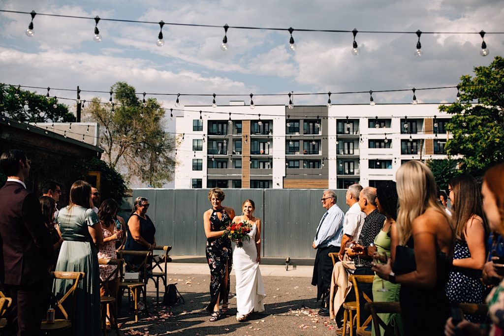 A bride walking down the aisle of her wedding with her mother, the bride is holding a flower bouquet, the wedding guests are all standing and watching at Blanc Denver