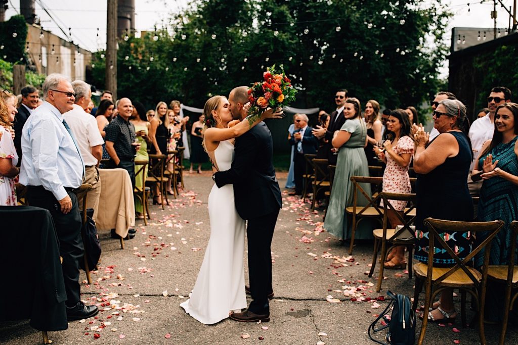 A bride and groom kiss after their intimate outdoor wedding reception at The Ramble Hotel in downtown Denver, the bride holds a flower bouquet behind the grooms head as their guests stand and clap on either side of the couple at Blanc Denver