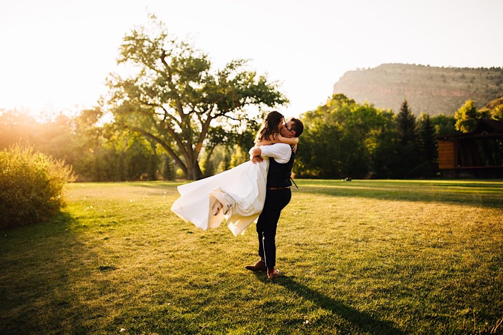 Bride and groom kiss as the groom holds and swings the bride through the air in a field at Planet Bluegrass after their wedding ceremony