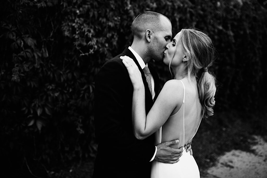 Black and white photo of a bride and groom kissing while wearing their wedding attire in front of a bush