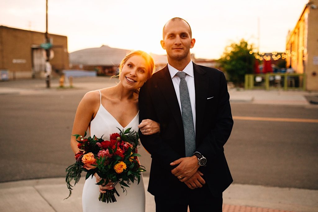 A bride and groom in wedding attire stand next to one another with their arms locked looking at the camera, both are smiling and the bride is holding a flower bouquet with the sun setting behind them in downtown Denver 