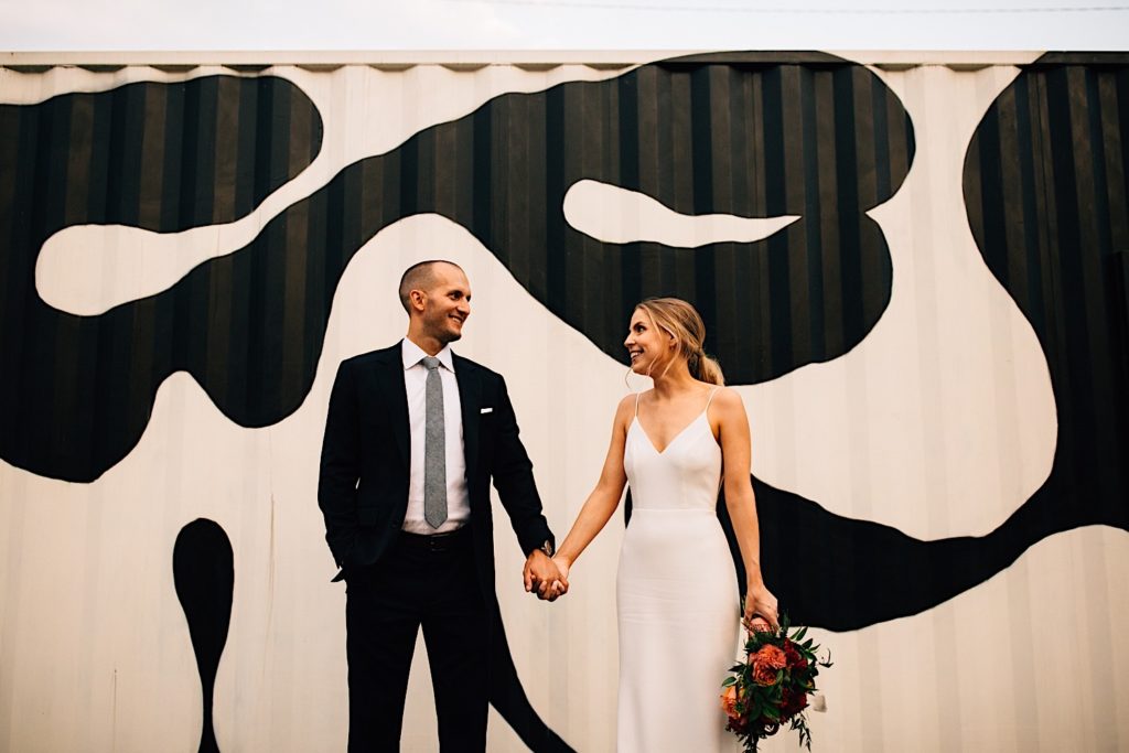 Bride and groom standing next to one another holding hands and smiling while looking at each other, the bride is holding a flower bouquet and they are in front of metal siding with a clack and white cow pattern on it