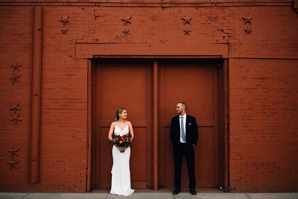 Bride and groom standing in their wedding attire in front of a orange/brown brick wall smiling at one another, there is a double doorway on the wall and they are each in front of one door near Blanc Denver