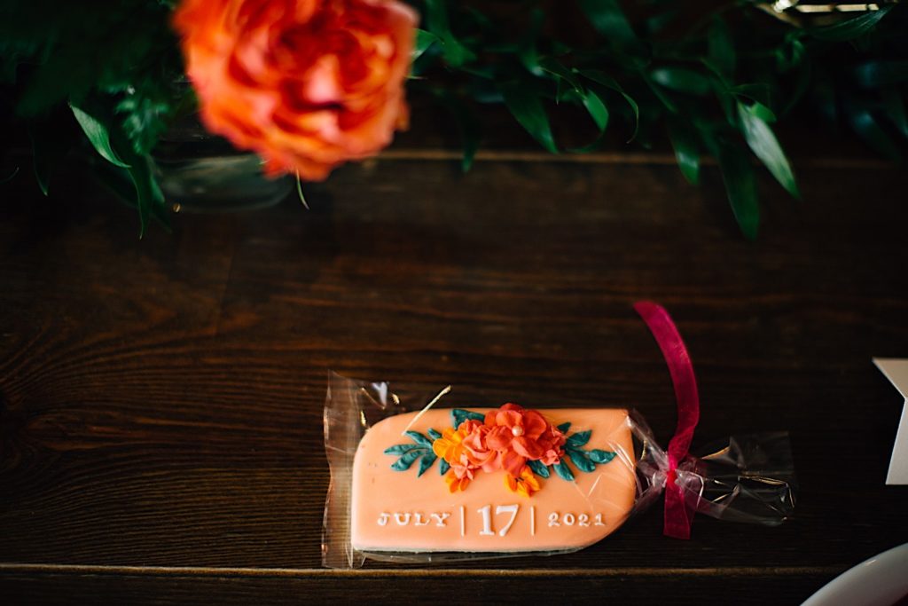 Close up of a wrapped cookie with frosting flowers and "July | 17 | 2021" written on it. The cookie is on a wooden table with a real flower behind it at Blanc Denver