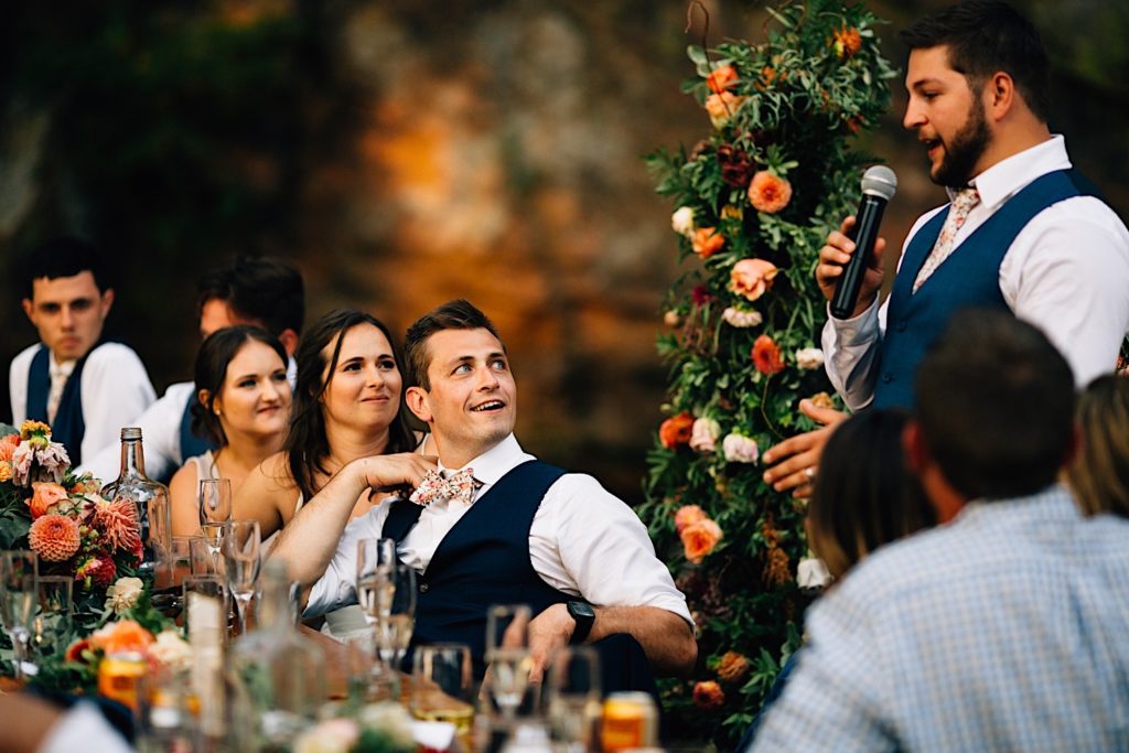 Bride and groom smile and watch as a groomsmen gives a speech during their wedding reception