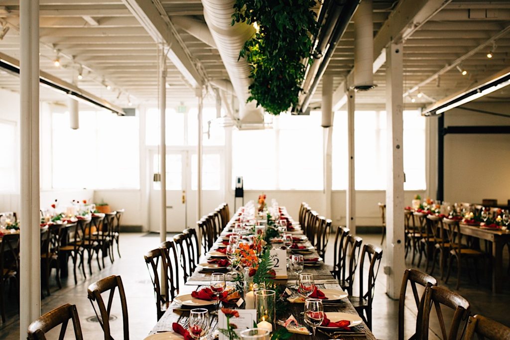 Table set for a wedding reception decorated with flowers and cookies. greenery hangs from the ceiling above the table and there are other tables on either side at Blanc Denver