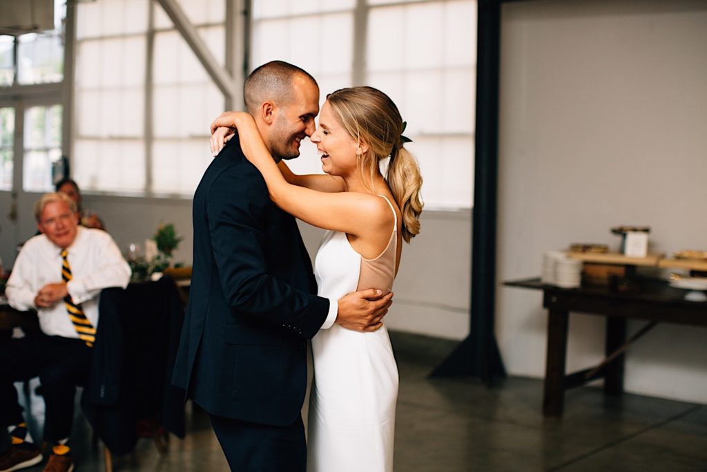 Bride and groom dancing with one another during their wedding reception and laughing, there are guests seated and watching behind the groom at Blanc Denver