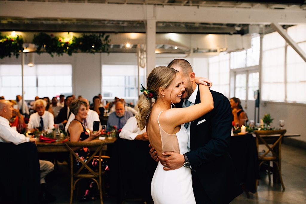 Bride and groom dancing with one another during their wedding reception and smiling, there are guests seated and watching behind them at Blanc Denver