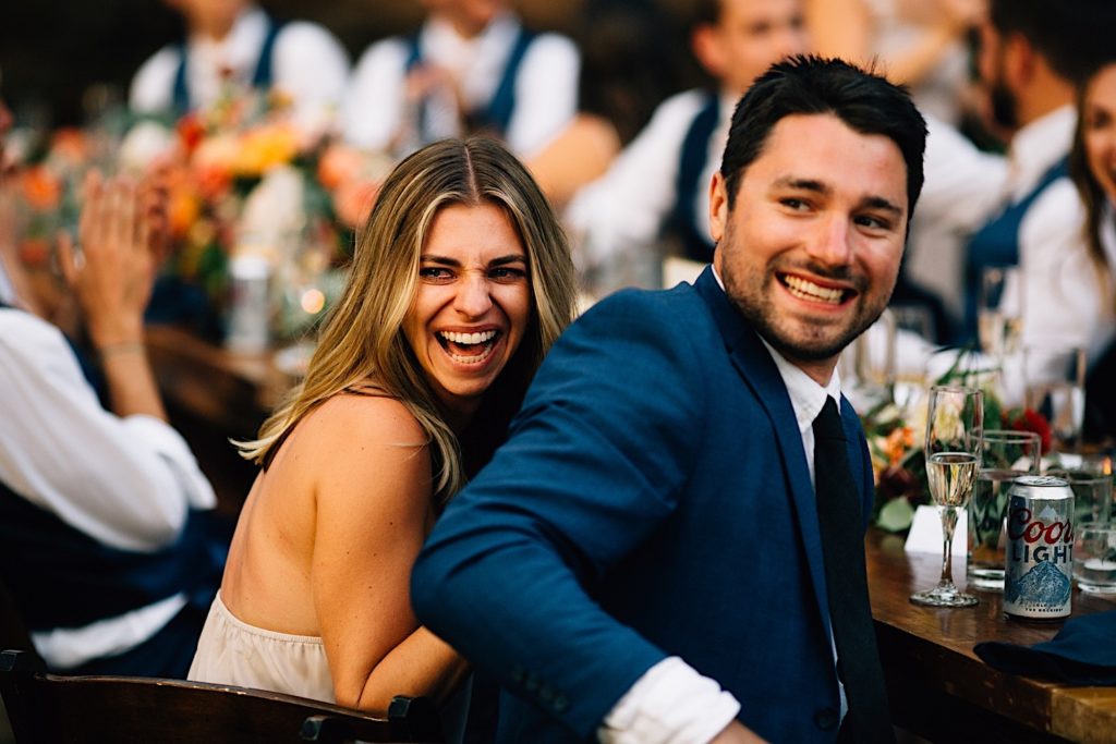 Guests seated at a wedding reception look behind them and laugh