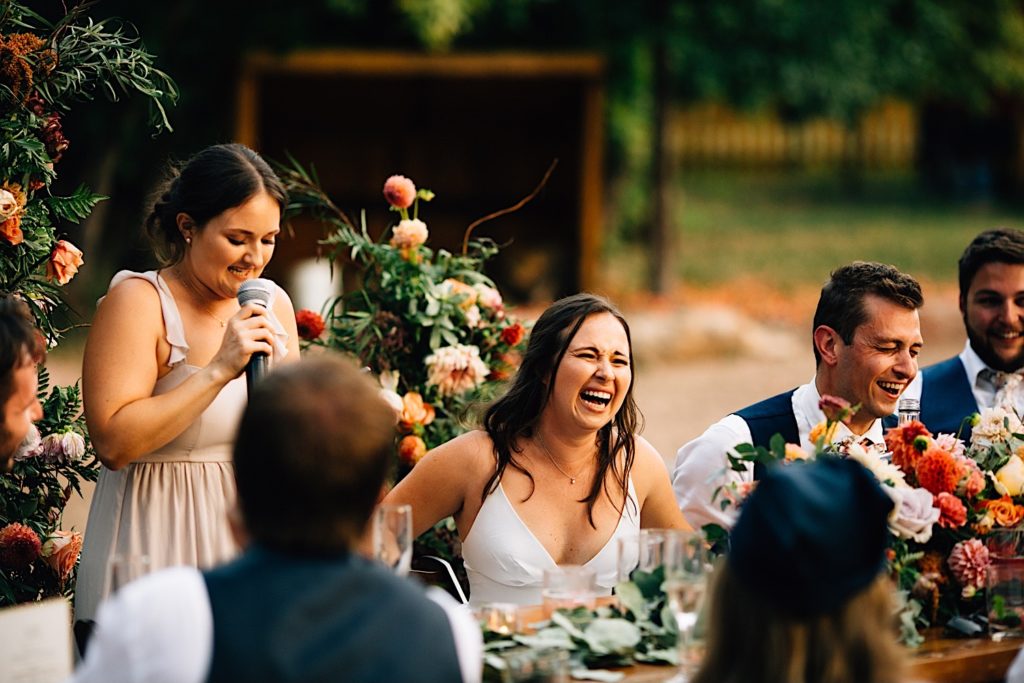 Bridesmaid giving a speech at a wedding reception at Planet Bluegrass while the bride and groom sitting next to her laugh