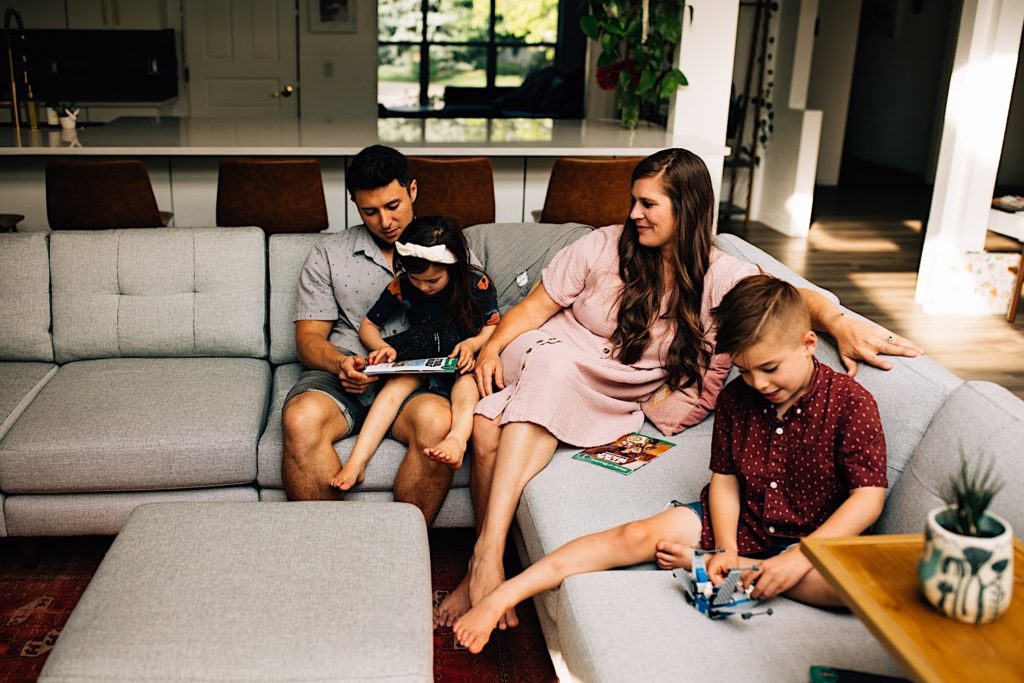 A family sits together on the couch, the father helps the daughter read while the mother smiles at them as her son plays with Legos next to her