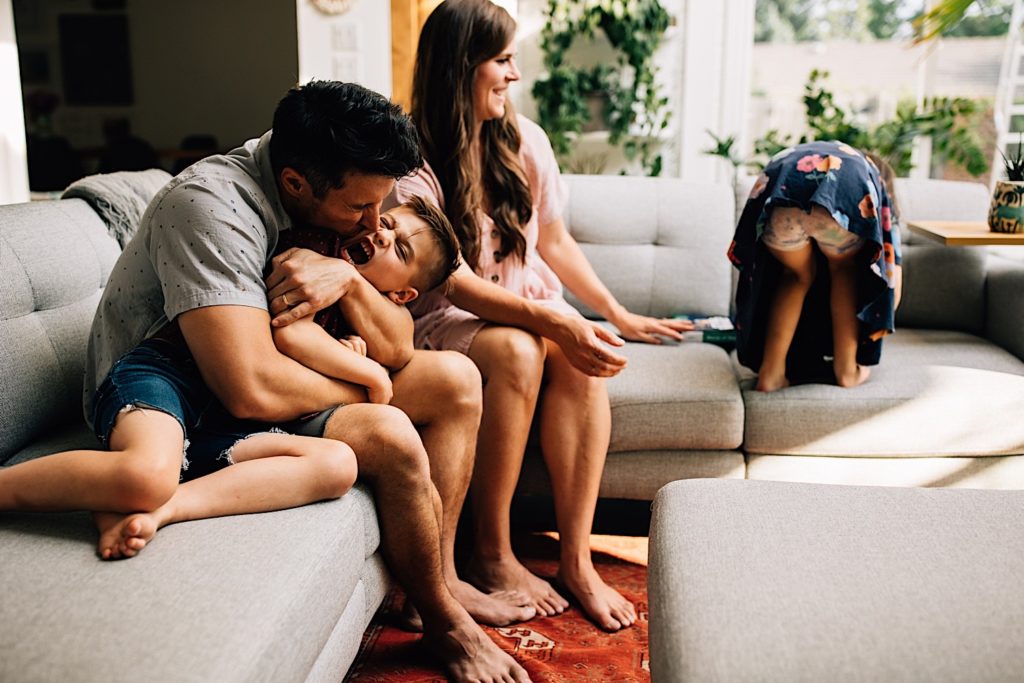 A family sits together on the couch, the father hugs and kisses his screaming son while the mother laughs at her daughter playing with toys on the couch