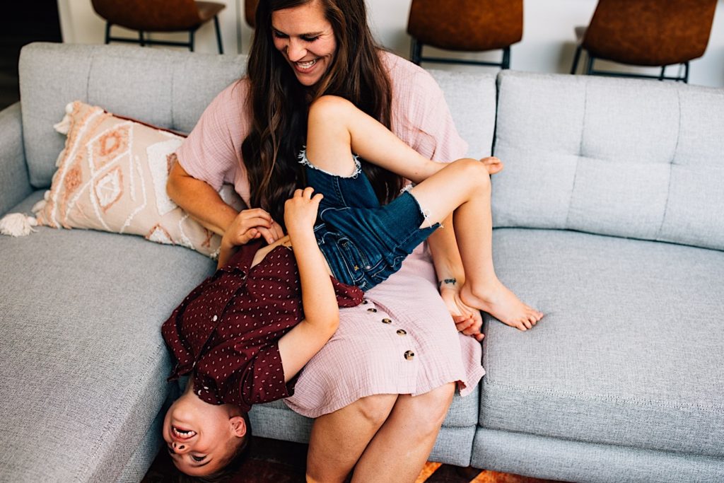 A mother sits on the couch laughing as she's tickling her laughing sun as he hangs upside down on her lap