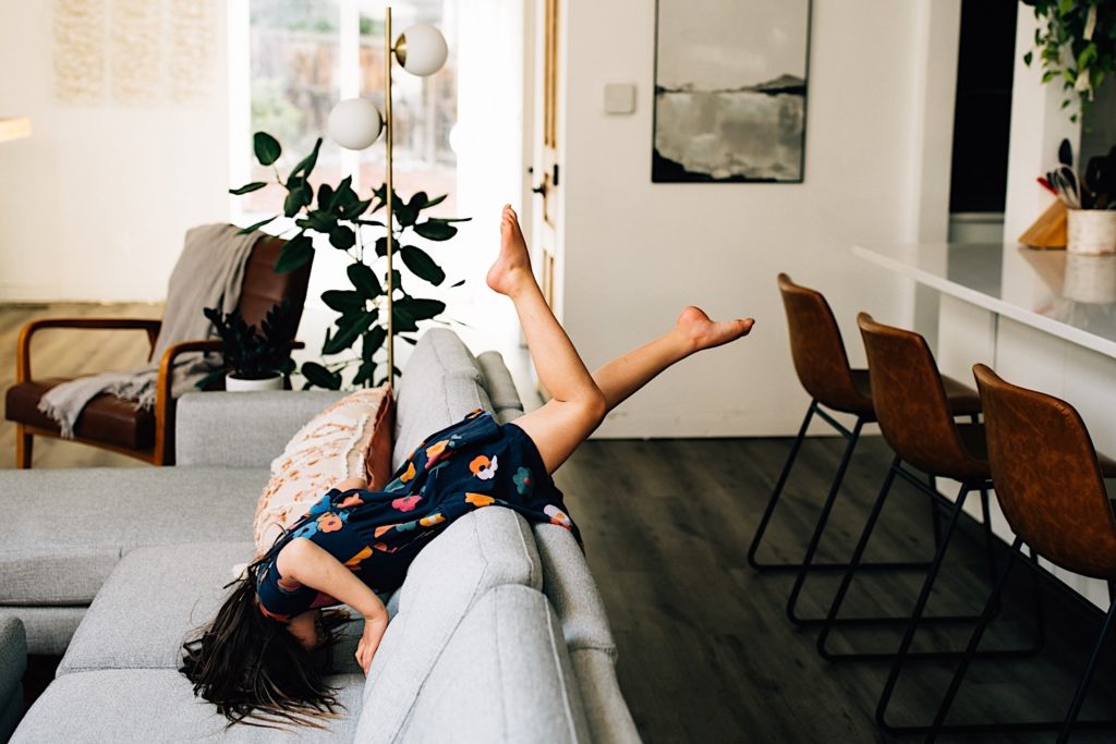 A child in a floral dress climbs over the back of a couch and tumbles head first down it