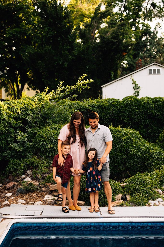 A family standing outside together in their backyard next to their pool with shrubs in the background. The kids are smiling at the camera while their parents look down at them.