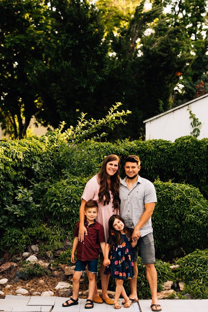 A family standing outside together in their backyard next to their pool with shrubs in the background smiling at the camera