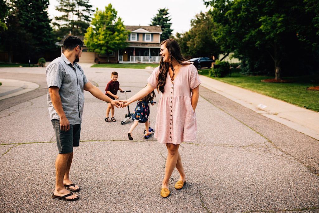 A mother and father stand in the street holding hands, the father is looking at his children on their scooters behind him while his wife smiles and looks at him.