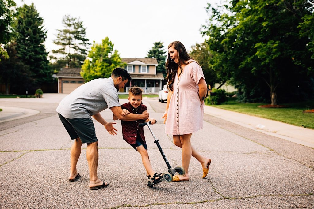 A family stands in the street together, the father catches his son as he rides his scooter between him and his wife.