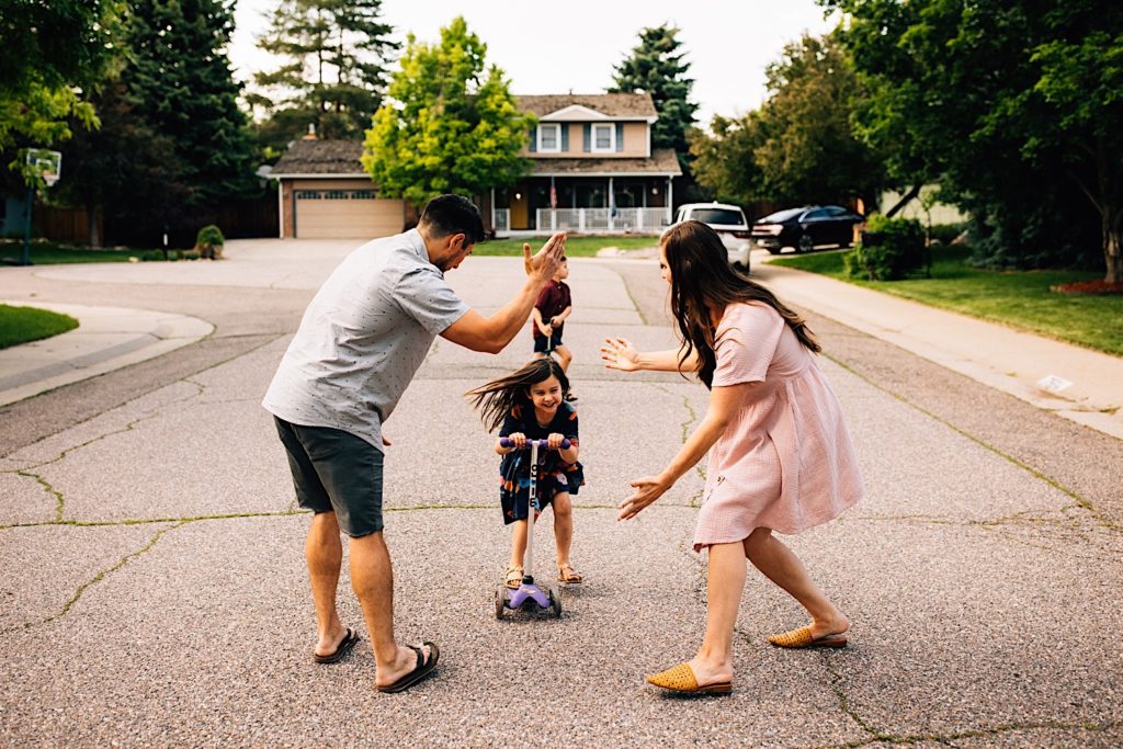 A family stands in the street together, the parents are looking at their daughter and lifting their hands in the air as she rides her scooter towards them while smiling.