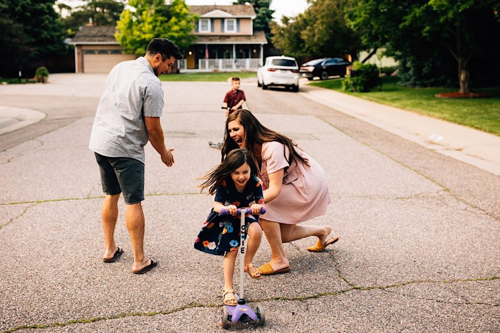 A family stands in the street together, the daughter just rode her scooter between her parents and is smiling while her mother is crouched and laughing behind her.