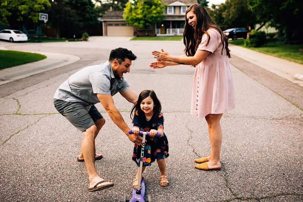 A family stands in the street together, the father catches his daughter as she rides her scooter between him and his wife as the wife claps