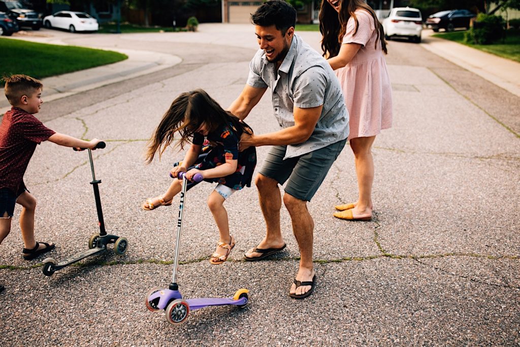 A family stands in the street together, the father catches his daughter as she rides her scooter between him and his wife as the wife claps, the son is walking his scooter towards them