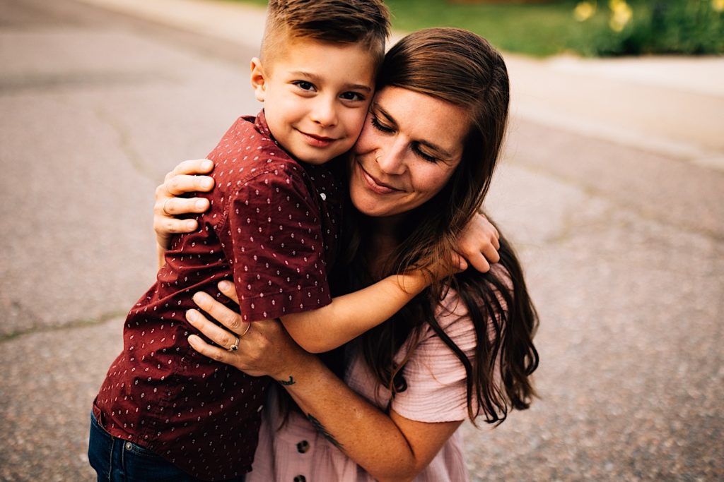 A mother and son hug in the street, the son is looking at the camera while the mother is crouched next to him with her eyes closed