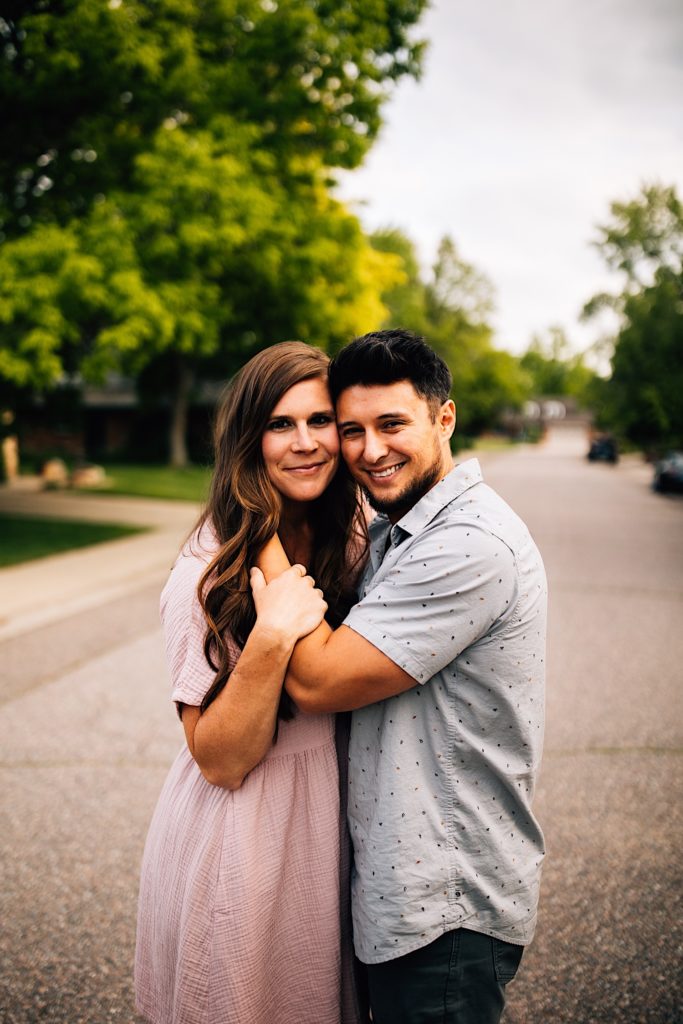 A couple stand together in the street, the man has his hand on the woman's face while she holds his arm as they both smile at the camera